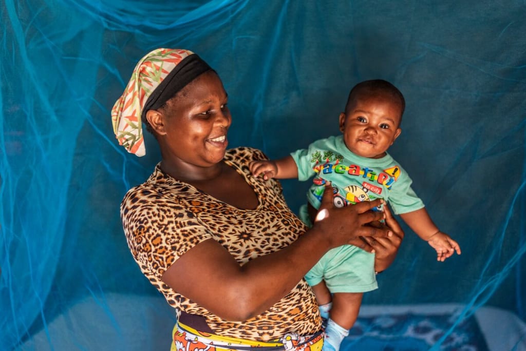Grace is wearing a brown and black animal print dress with a yellow patterned skirt. She is holding Emmanuel and smiling at him. Emmanuel is looking at the camera and smiling. They are standing in front of a bed that is covered with a mosquito net.